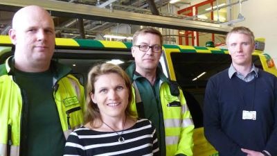 Tre män och en kvinna står och ler framför en ambulans inne på en brandstation.