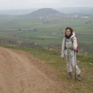 Santiago de Compostelan pyhiinvaellusreitillä tien reunassa avarassa maisemassa taustalla matalaa vuoristoa, Manuela Bosco seisoo vaellussauvaan nojaten ja katsoo kameraan.