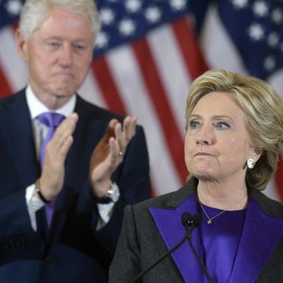 Hillary Clinton katsoo kuvan vasempaan laitaan vakavana. Taustalla taputtaa Bill Clinton. Taustalla on myös Yhdysvaltain lippuja.