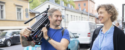 A photograph in a blue shirt carries a camera tripod and talks to a journalist in a blue shirt.