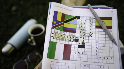 En tidning är vikt så att en sida med ett korsord syns. En penna vilar på tidningen och korsordet är delvis ifyllt. Orden Namibia, Italia, Spania, Tanzania och Rumänia är skrivna.