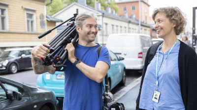 A photograph in a blue shirt carries a camera tripod and talks to a journalist in a blue shirt.