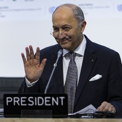 Ranskan ulkoministeri Laurent Fabius Pariisin ilmastokokouksessa. Tummaan pukuun ja pilkulliseen kravattiin pukeutunut Fabius hymyilee. Hänen silmälasinsa ovat matalalla nenän päällä ja hän pitää oikeaa kättään hieman ylhäällä olkapään tasolla, kämmen kameraan päin. Ulkoministerin edessä pöydällä on mikrofoni. Tausta on valkoinen, siinä näkyy ilmastokokouksen vihreitä logoja.