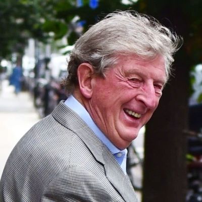 En skrattande Roy Hodgson i grå kavaj promenerar på en gata.