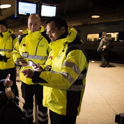 Kirkkaankeltaiseen vartijan asuun pukeutunut mies tarkistaa vaalean sinitakkisen naisen henkilöllisyystodistusta. Taustalla näkyy kaksi muuta samanlaisiin keltaisiin pusakoihin pukeutunutta miestä.