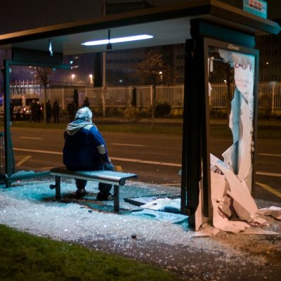 Mies istuu bussikatoksessa, lasiseinät sirpaleina kadulla