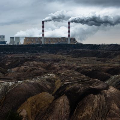 Voimalan piipuista tupruaa taivaalle paksusti savua. Kuvan etualalla kasvillisuudesta paljasta kalliota.