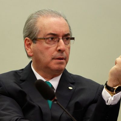 Eduardo Cunha istuu mikrofonin takana ja pitää vasenta kättään pystyssä.