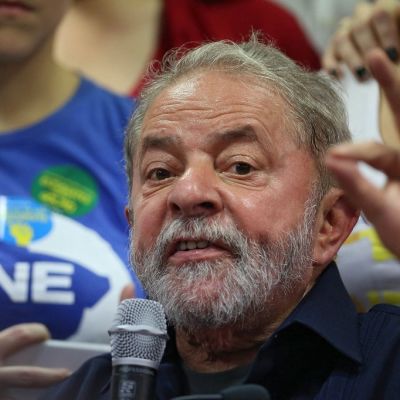 Parrakas Lula da Silva puhuu oikeassa kädessään olevaan mikrofoniin ja elehtii vasemmalla kädellään, jonka etusormi ja peukalo ovat yhdessä. Lulalla on tummansininen kauluspaita. Taustalla seisoo kannattajia.