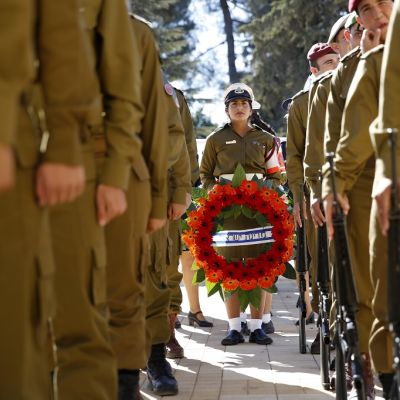 Israelilainen naissotilas pitelee punaisista kukista tehtyä seppelettä. Molemmilla puolilla rivi sotilaita kivääreineen.