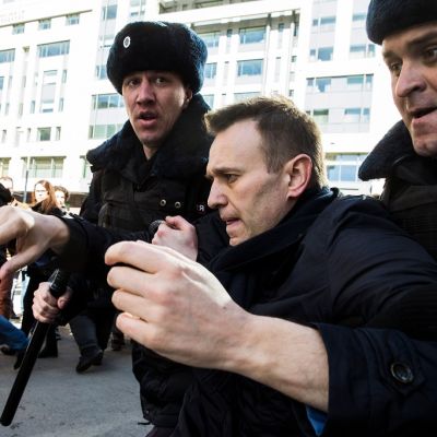 Kaksi poliisimiestä karvalakeissa kuljettaa Navalnyia kainaloista nostaen. Taustalla näkyy ihmisjoukkoa ja auringon kirkkaasti valaisema kerrostalon seinä. Navalnyi on paljan päin, mustassa takissa.