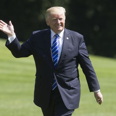 Donald Trump kävelee Valkoisen talon nurmikolla tummassa puvussa, sinisessä valkoraitaisessa kravatissa, viittoo oikealla kädellään ja hymyilee.