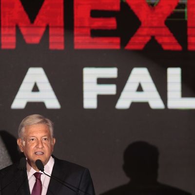 López Obrador seisoo tummassa puvussa ja punaisessa kravatissa lavalla mikrofonin takana. Hänen takanaan mustalla pohjalla lukee isoin punaisin kirjaimin "Mexico".