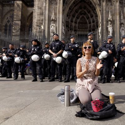 Hymyilevä, käsiään taputtava nainen polvillaan Kölnin tuomiokirkon edessä. Maassa reppu, termospullo ja eväspussi. Takana rivi poliiseja. 