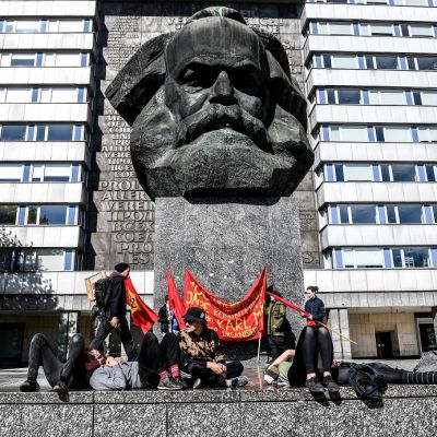 Pieni joukko nuoria ihmisiä punaisten banderollien kanssa makailee ja istuskelee jyhkeän betonisen Marxin päätä esittävän patsaan jalustan juurella. Taustalla vaalea DDR-tyylinen kerrostalo.