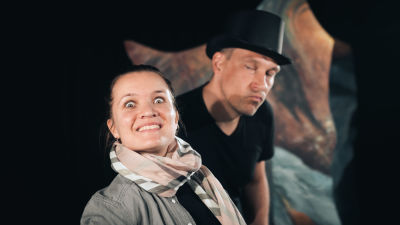 En kvinna och en man står på en teaterscen och ser in i kameran. Mannen har en hög hatt på huvudet.