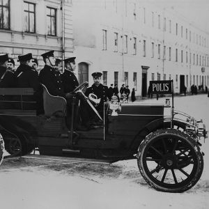 Ett gammalt polisfordon från början av 1900-talet fullastat med poliser på en snöig gata i en stad med höghus.