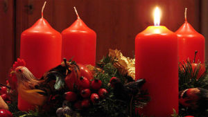 Neljä paksua punaista kynttilää, joista yksi sytytetty, jouluisessa koristekranssissa.