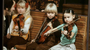 Linda Lampenius (till vänster) i TV-programmet Viuluviikarit musiikkimmaassa under ledning av Geza Szilvay Barn sitter på en bänk och spelar violin