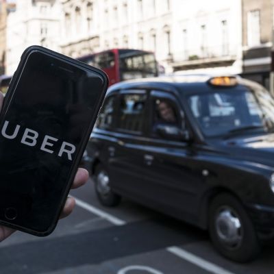 Etualalla näkyy älykännykkä, jonka näytöllä lukee isoilla kirjaimilla UBER. Taustalla näkyy musta Lontoon taksi.