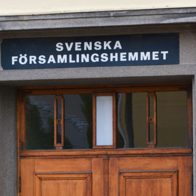 Närbild på en gul byggnad. I mitten av bilden syns övre delen av en trädörr. Ovanför den står det Svenska församlingshemmet med versaler.