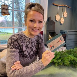 Närbild av en leende kvinna med uppsatt hår som håller i ett handgjort julkort. Kvinnan är Lee Esselström, programledare för livsstilsprogrammet Strömsö.