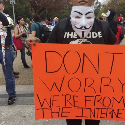Guy Fawkes -naamiolla varustautunut mielenosoittaja pitelee kylttiä, jossa lukee "Älä huoli, olemme internetistä".