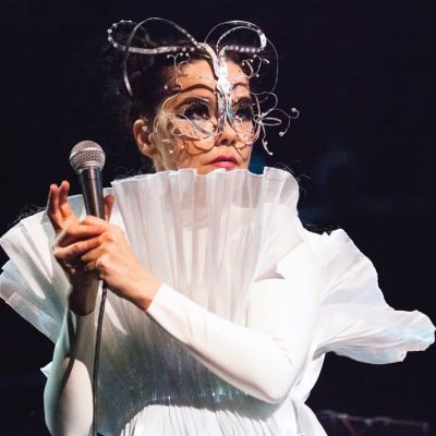 Artisten Björk med mikrofon i handen på scen i vita kläder och konstiga smycken i ansiktet.