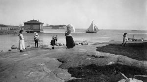 Förr var det var vanligt att skydda sig från solen med täckande kläder och parasoll. Fotot taget i Brunnsparken i Helsingfors år 1898. Fotograf okänd. Människor vid stranden.