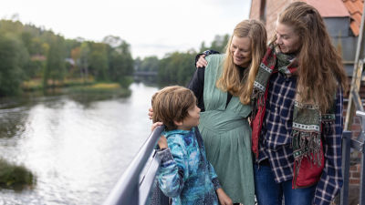 Två kvinnor står nära varandra tillsammans med en pojke. De står vid en å.