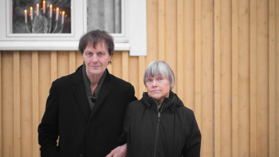 Två personer står framför ett gult trähus. De ser in i kameran med allvarliga miner.