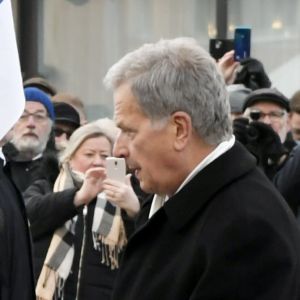 Sauli Niinistö håller i en blåvit banderoll som är fäst vid en blomkrans, omringad av människor, på Kaserntorget i Helsingfors.