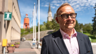 En medelålders man iklädd kostym, ljusröd skjorta och glasögon står på gatan i centrala Hangö, i bakgrunden vattentornet