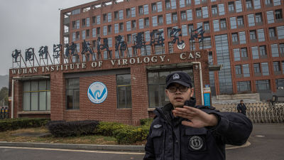 Suuri tiiliseinäinen rakennus, jossa lukee Wuhan Institute Of Virology. Vartija koittaa kedällään estää kuvaajaa ottamasta kuvaa rakennuksesta.
