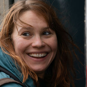 Leveästi hymyilevä nuori nainen (näyttelijä Seidi Haarla) on astumassa linja-auton tai paikallisjunan ovesta sisään reppu selässään ja katsoo taakseen, kuva elokuvasta Hytti nro 6.