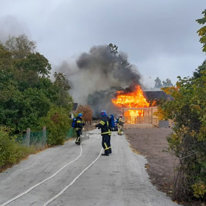 Ett hus brinner i bakgrunden, brandmän på väg att släcka