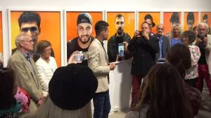 Ahmed Alalousi & Stefan Bremer. Vernissage på Galleri Elverket i Ekenäs. Opening of the exhibition at Galleri Elverket in Ekenäs. Vernissage av en fotoutställning. Färggranna foton hänger på väggen. Två fotografer i förgrunden, den ena fotar den andra med sin telefon. Publik syns nere på bilden.