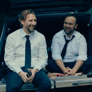 Två skrattande män sitter på kanten till en likbil.