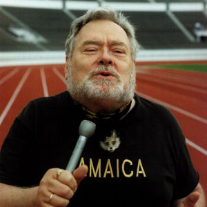 Jamaica-paitainen toimittaja Paavo Noponen stadionin juoksuradalla mikrofoni kädessään.