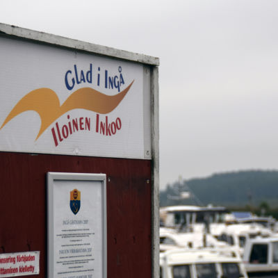 En skylt där det står Glad i Ingå på svenska och finska. I bakgrunden syns båtar i en småbåtshamn,