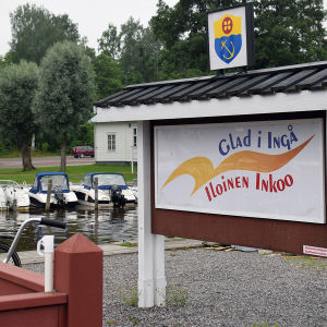 En skylt där det står Glad i Ingå på svenska och finska. I bakgrunden syns en småbåtshamn.