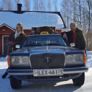 Kaksi miestä nojaavat taksiin lumisella pihalla.