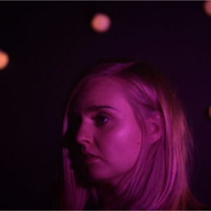 Vaaleahiuksisen naisen pää keskellä pimeyttä, muutama valo näkyy taustalla. Kasvoihin heijastuu violetti valo.