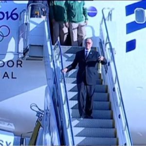 Silmälasipäinen mies tummassa puvussa laskeutuu lentokoneesta portaita alas lyhdynnäköinen kullankeltainen esine kädessään. Koneen kyljessä on olympiarenkaat ja siinä lukee Rio 2016, Apoiador oficial.