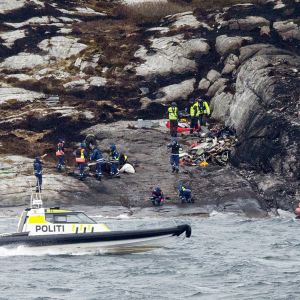 Joukko poliiseja ja pelastustyöntekijöitä tutkii rantakallioita. Saaren edustalla ajaa poliisin moottorivene. Oikeassa reunassa punainen kumivene ajaa kohti rantaa.