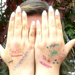 En flicka som håller händerna för ansiktet. På händerna står skrivet olika komplimanger så som du är vacker.