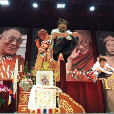 Taistalla Dalai laman kasvoja kirjojen kansissa. Edessä tanssija hyppää korkealle ilmaan. 
