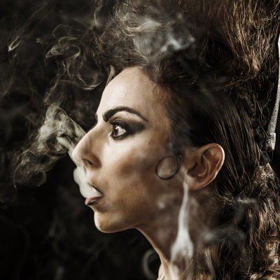 Tummatukkainen nainen puhaltaa tupakan savua suustaan.