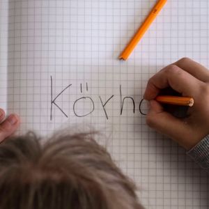 Kuvassa on lapsen kädet ja pää kumartuneena vihkon ylle. Lapsi kirjoittaa ruutuvihkoon sanaa köyhä.