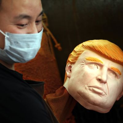 Mies hymyilee, katsoo alaviistoon hengityssuojain suun edessä, kädessään Trumpin kasvoja esittävä kuminaamari.